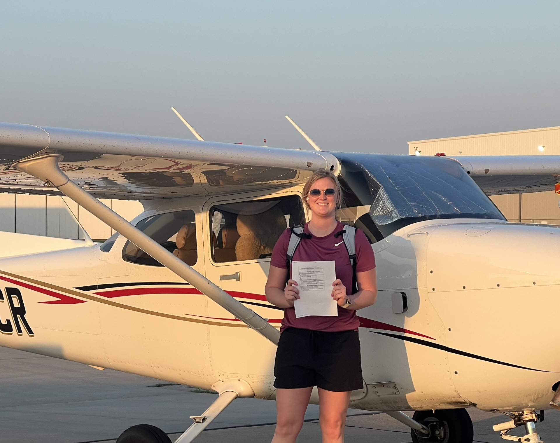 Smiling pilot holding her certificate in front of a single-engine aircraft on the ramp in warm evening light