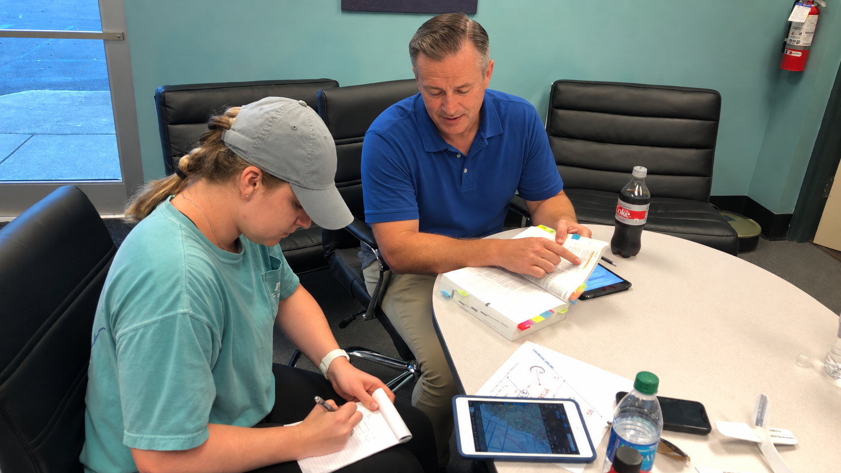 Instructor coaching a student through a tabbed pilot manual at a training table