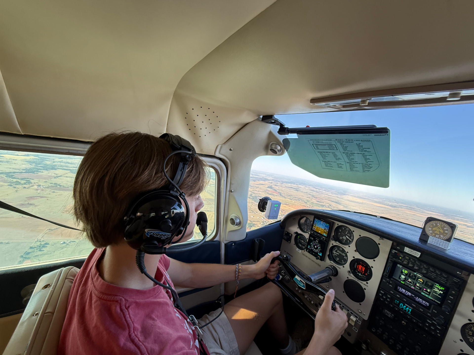 Young pilot at the controls wearing a headset, flying a light aircraft over open farmland
